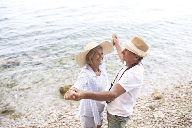 Активная милая пожилая пара, держащаяся за руки и танцующая на пляже — стоковое фото