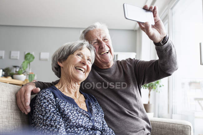 Lachende Senioren Sitzen Auf Der Couch Im Wohnzimmer Und Machen Selfie Mit Dem Smartphone Innenraume Zuversicht Stock Photo