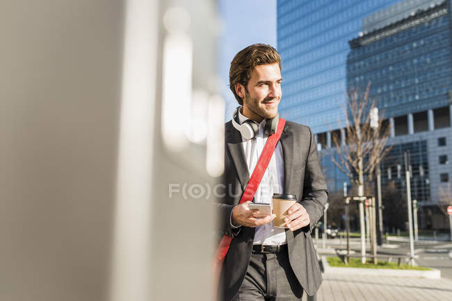 Молодой бизнесмен гуляет по городу с чашкой кофе, используя мобильный телефон — стоковое фото