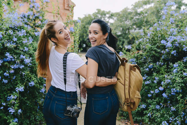 Two happy young women with camera and backpack embracing walking among blooming bushes — Stock Photo