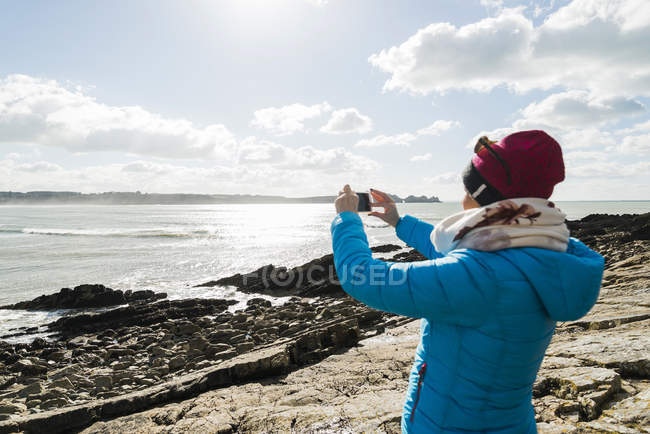 Mujer de pie en la costa rocosa tomando una foto, Francia, Bretaña, Finistere, península de Crozon - foto de stock