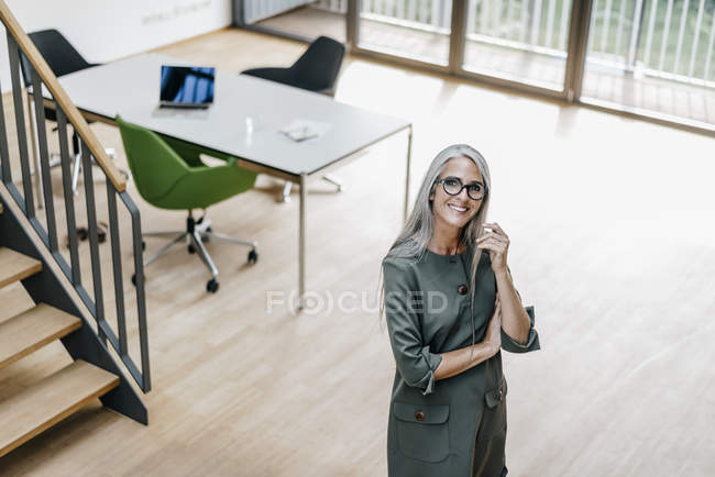 Retrato de una mujer sonriente con el pelo largo y gris de pie en la oficina - foto de stock