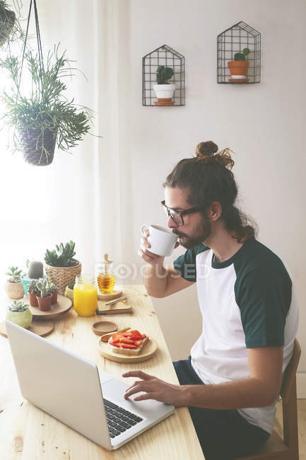 Hombre joven usando el ordenador portátil mientras desayuna - foto de stock