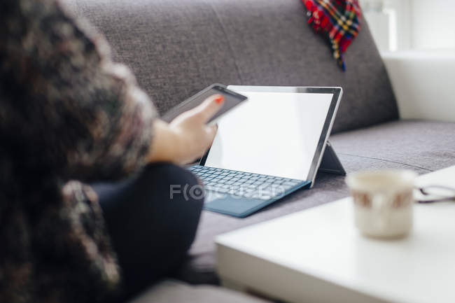 Young woman sitting on the couch using tablet and smartphone — Stock Photo