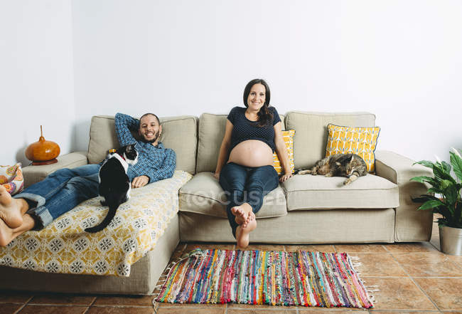 Felice coppia in attesa di un bambino seduto sul divano a casa con due gatti — Foto stock