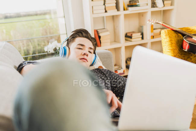 Junger Mann liegt mit Laptop und Kopfhörer auf Couch — Stockfoto