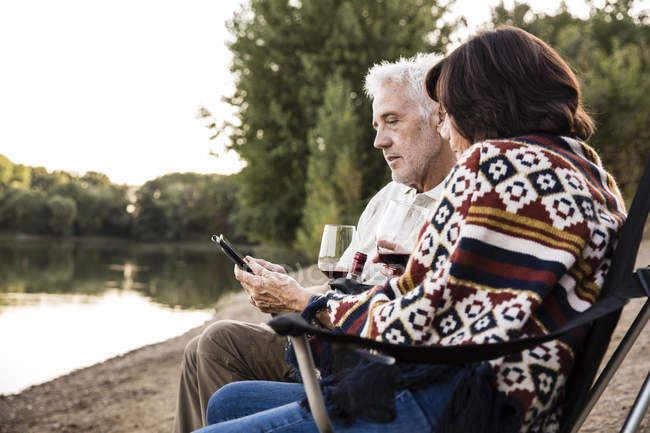 Senior couple with wine sitting at a lake with tablet — Stock Photo
