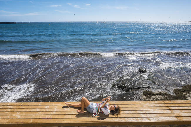 Frau liegt auf Bank vor dem Meer und benutzt Smartphone, Teneriffa, Spanien — Stockfoto