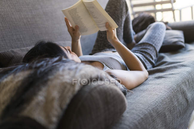 Mujer joven acostada en el sofá, leyendo el libro - foto de stock