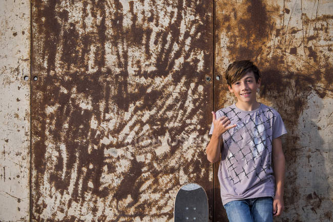 Портрет мальчика, прислонившегося к ржавой металлической стене со скейтбордом — стоковое фото