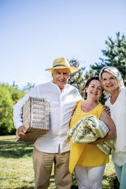 Ritratto di amici anziani sorridenti in piedi su un prato con coperta e cestino da picnic — Foto stock