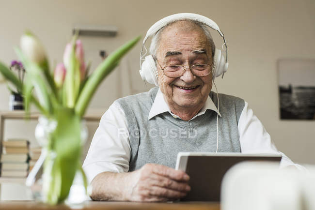 Hombre mayor usando mini tableta y auriculares en casa - foto de stock