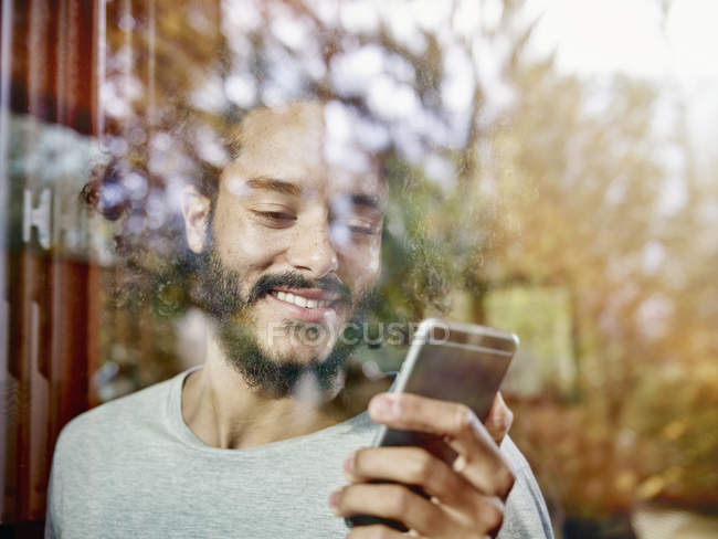 Smiling young man looking on cell phone behind windowpane — Stock Photo