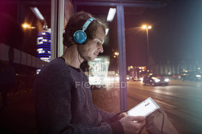 Uomo con cuffie seduto alla fermata dell'autobus utilizzando tablet digitale di notte — Foto stock