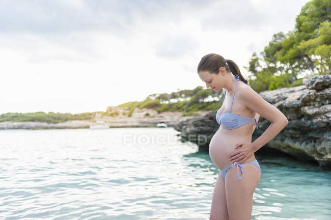 Беременная женщина, стоящая в море на скалистом побережье — стоковое фото