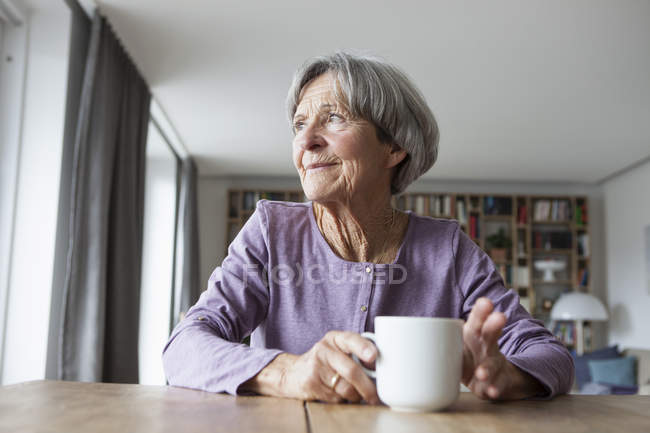 Portrait of senior woman sitting at table with cup of coffee looking through window — Stock Photo