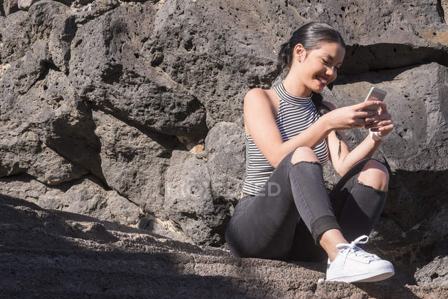 Молода жінка сидить перед кам'яним обличчям, дивлячись на її смартфон — стокове фото