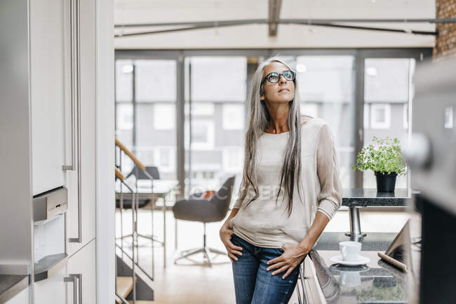 Femme avec de longs cheveux gris debout dans la cuisine moderne et regardant vers le haut — Photo de stock