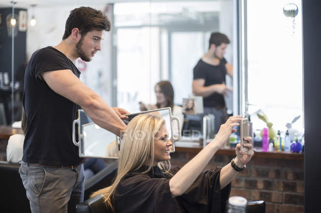 Mujer en peluquería revisando el peinado en el espejo y tomando selfie —  conexión, Belleza - Stock Photo | #177235134