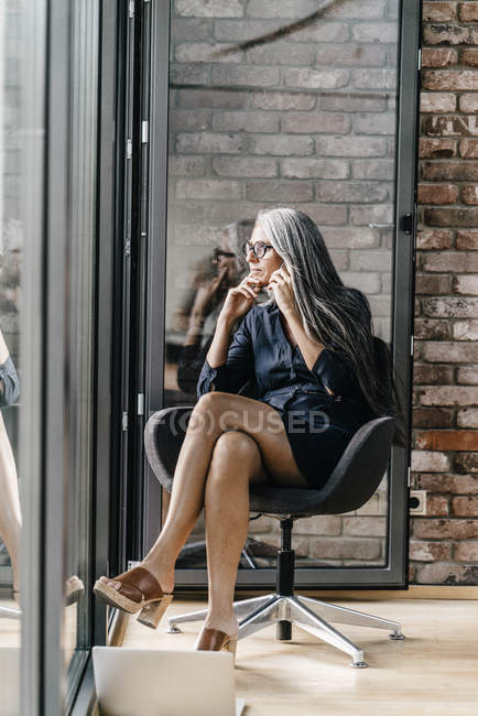 Frau mit langen grauen Haaren am Handy sitzt am Fenster — Stockfoto