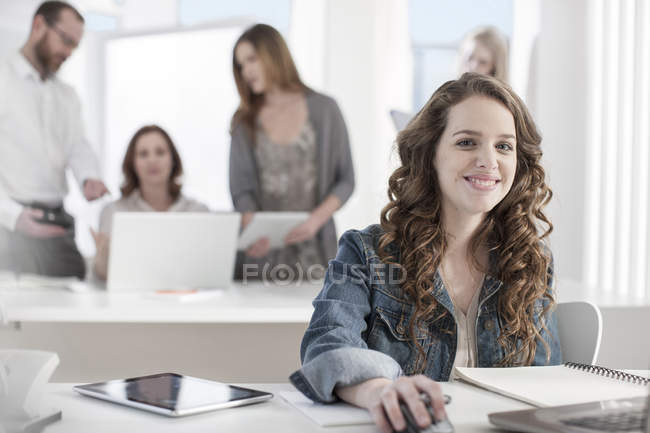 Портрет улыбающейся женщины, сидящей за столом в офисе с коллегами на заднем плане — стоковое фото