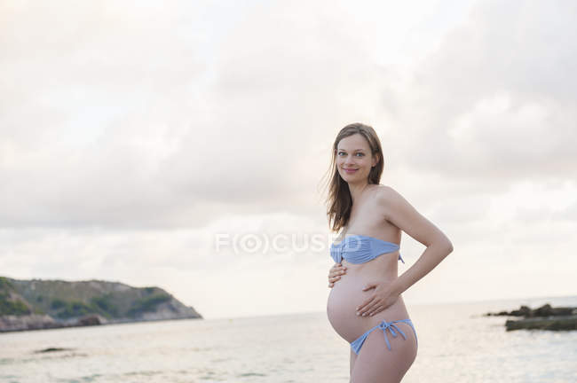 Sorrindo mulher grávida de pé na praia e olhando para a câmera — Fotografia de Stock