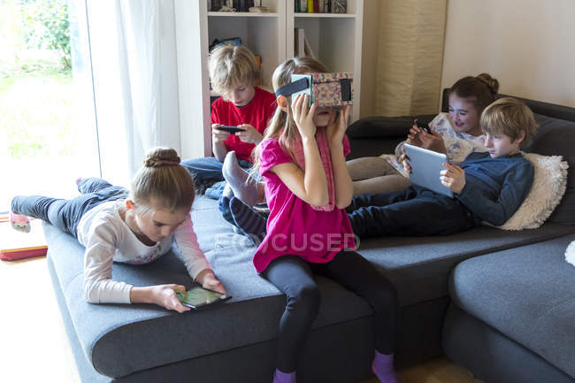 Fünf Kinder auf einer Couch mit verschiedenen digitalen Geräten — Stockfoto