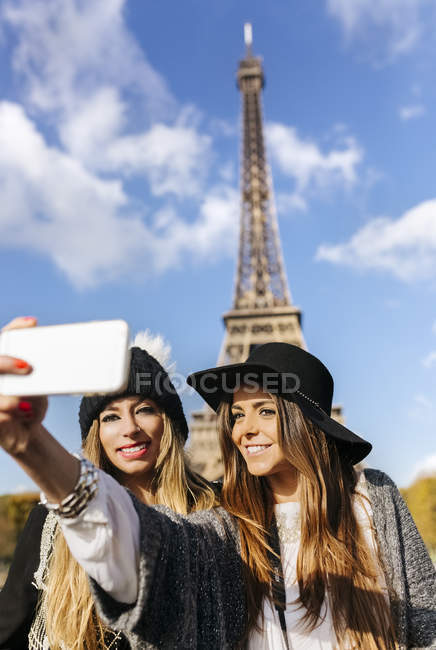 Francia, París, dos mujeres sonrientes tomando una selfie con la Torre Eiffel en el fondo - foto de stock