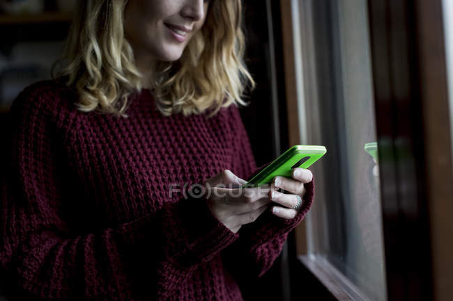 Улыбающаяся женщина стоит у окна и смотрит на смартфон. — стоковое фото
