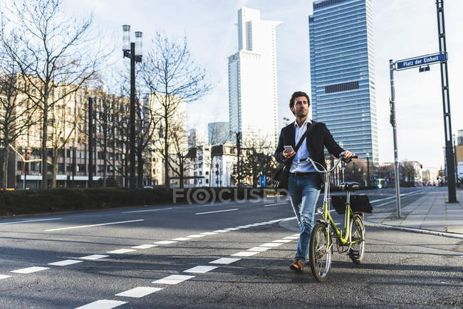 Germany, Frankfurt, Young businessman in the city with bicycle, using mobile phone — Stock Photo