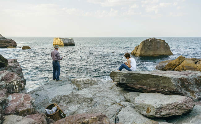 Hombre mayor pescando en el mar con su esposa sentada en la roca - foto de stock