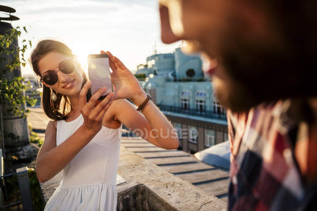 Jeune femme souriante prenant une photo de son petit ami sur une terrasse sur le toit — Photo de stock