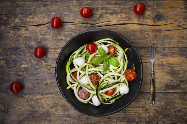 Bowl of zucchini spaghetti with mozzarella, cherry tomatoes and basil on wood — Stock Photo
