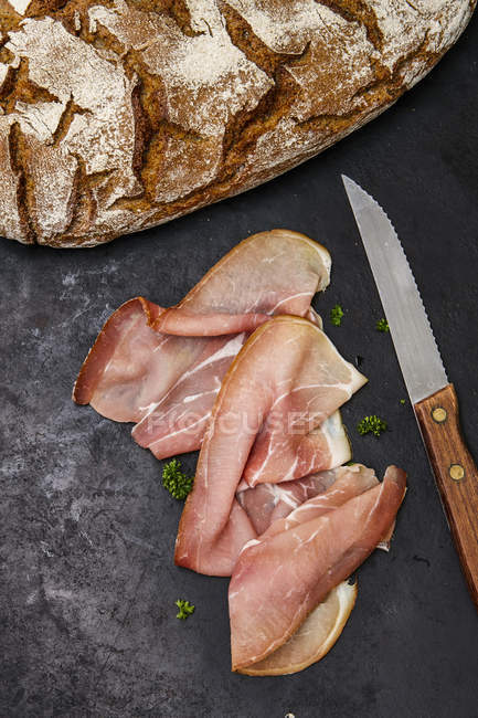 Slices of raw ham, parlsey, knife and bred on black surface — Stock Photo
