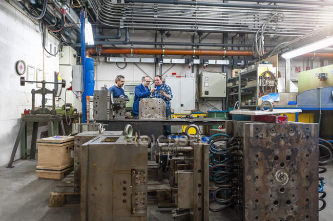 Three men in factory talking at metal workpiece — Stock Photo