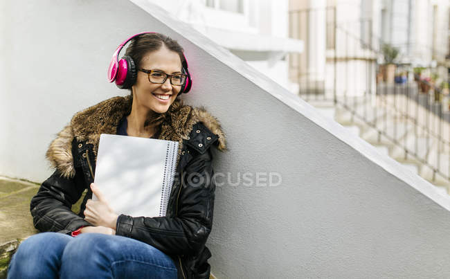 Student girl with headphones and writing pad leaning against building wall — Stock Photo