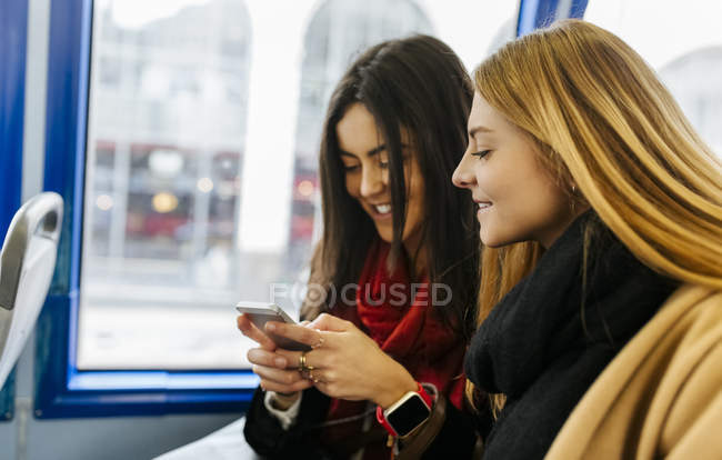 Dos mujeres jóvenes usando el teléfono inteligente en el autobús, Londres, Reino Unido - foto de stock