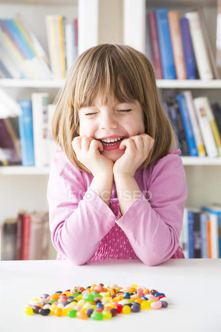 Retrato de niña sonriente con los ojos cerrados y gominolas en una mesa - foto de stock