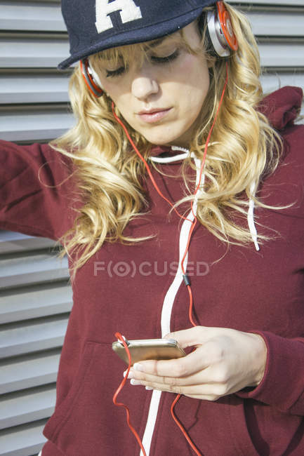 Блондинка в кепке и куртке в капюшоне слушает музыку со своего смартфона — 30 40 лет, день - Stock Photo