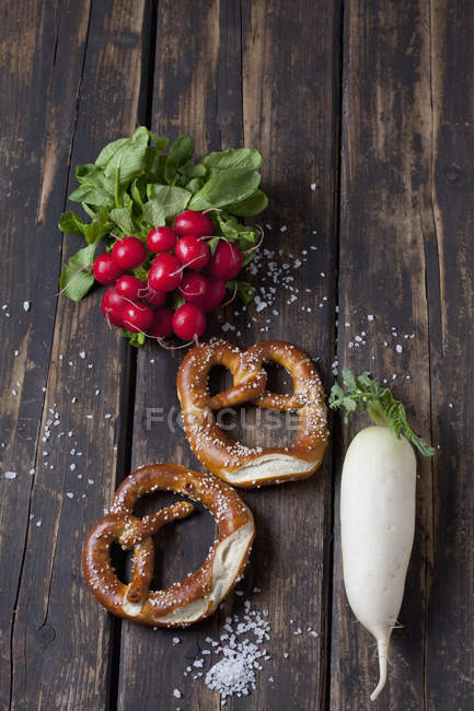 Two pretzels, red radishes, radish and scattered salt grains on dark wood — Stock Photo