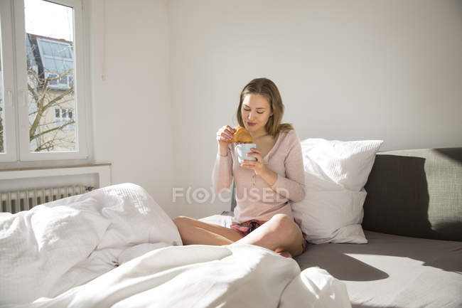 Jeune femme assise sur son lit avec une tasse de café et croissant — Photo de stock
