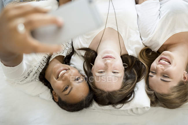 Três adolescentes felizes deitados na cama tirando uma selfie — Fotografia de Stock