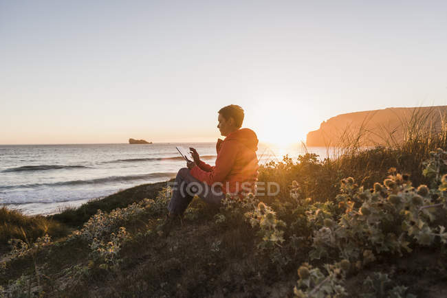 Mulher sentada na costa ao pôr do sol e usando tablet — Fotografia de Stock