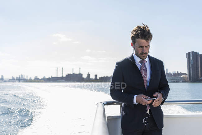 Uomo d'affari in piedi sul traghetto su East River con cellulare e auricolari, New York, Stati Uniti — Foto stock