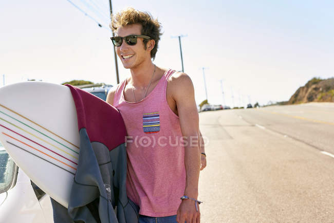 Jovem sorridente carregando prancha na estrada costeira — Fotografia de Stock