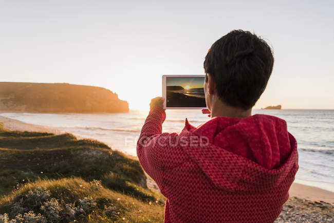 Femme prenant une photo avec un comprimé à la côte au coucher du soleil — Photo de stock