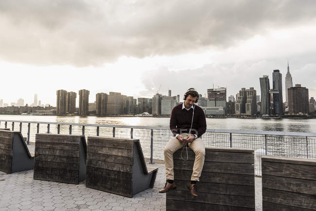Jovem com fones de ouvido e telefone celular sentado em East River, Nova York, EUA — Fotografia de Stock