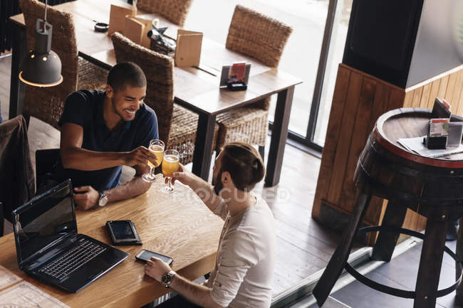Dois homens com laptop e dispositivos portáteis em um bar que clica copos de cerveja — Fotografia de Stock