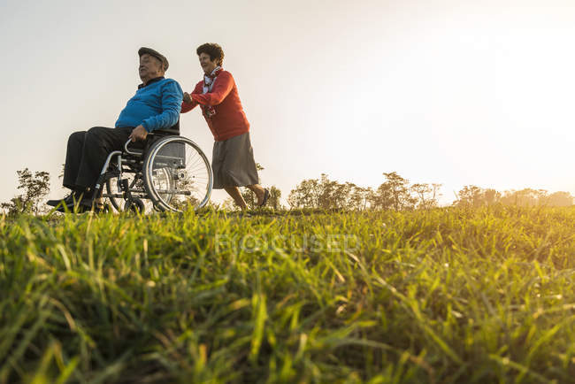 Старший жінка, штовхаючи чоловіка в інвалідному візку в природі — стокове фото