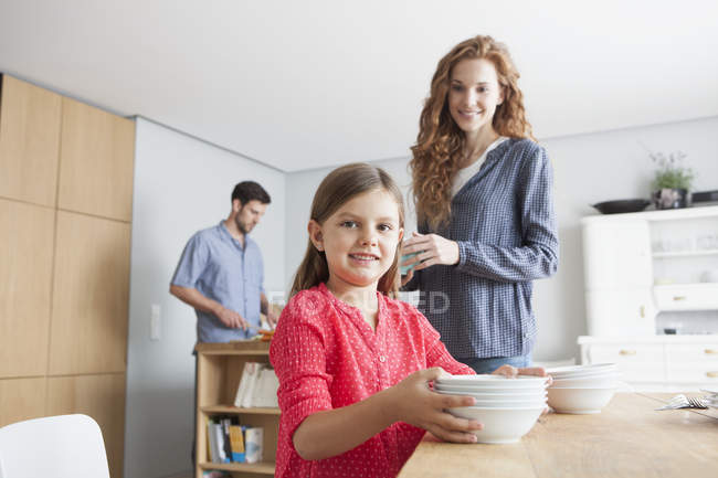 Portrait of little girl laying the table in the kitchen with her parents in the background — Stock Photo